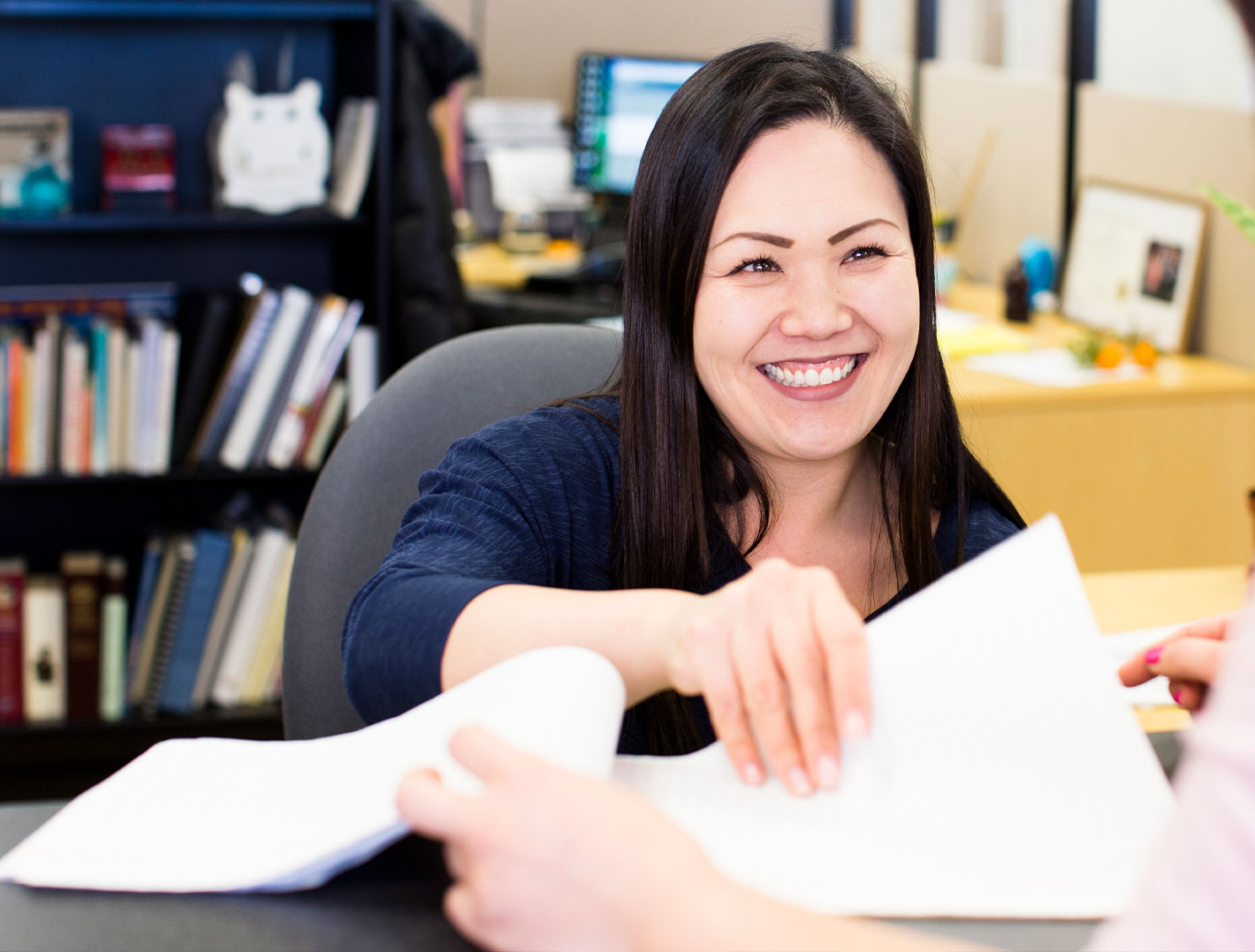 Smiling woman sitting at a help desk flipping through a stack of paper.